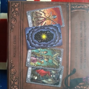 May include: A close-up of a tarot deck box with a third edition label. The box is decorated with ornate gold trim and features images of tarot cards, including the Seven of Wands, The Lovers, and The Devil.