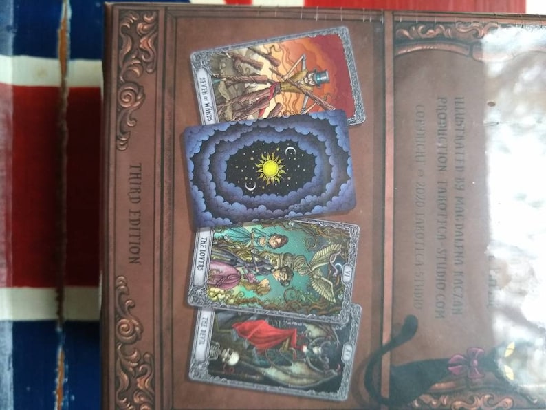 May include: A close-up of a tarot deck box with a third edition label. The box is decorated with ornate gold trim and features images of tarot cards, including the Seven of Wands, The Lovers, and The Devil.