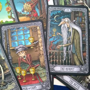 May include: A set of tarot cards with black borders and silver accents. The cards feature illustrations of people and scenes in a dark, gothic style. The cards include "The Hermit", "The Devil", and "Four of Cups".
