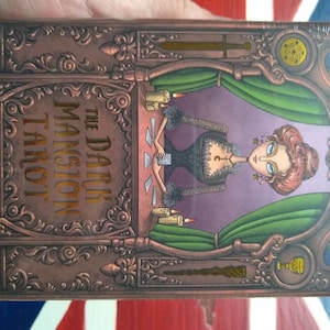 May include: The Dark Mansion Tarot deck box features an ornate, copper-colored design with a woman in a black dress and a green curtain behind her. The text on the box reads "The Dark Mansion Tarot".