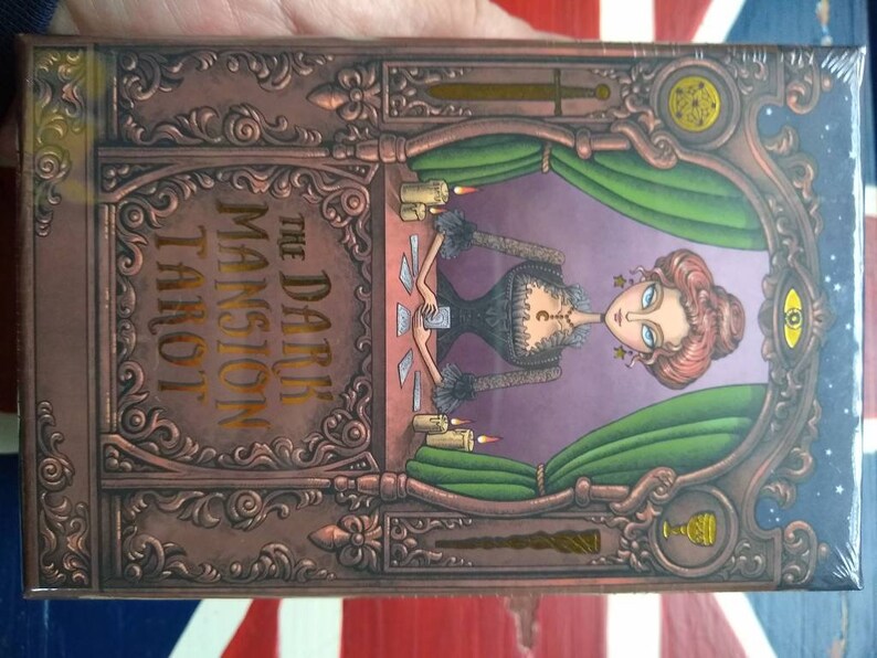 May include: The Dark Mansion Tarot deck box features an ornate, copper-colored design with a woman in a black dress and a green curtain behind her. The text on the box reads "The Dark Mansion Tarot".
