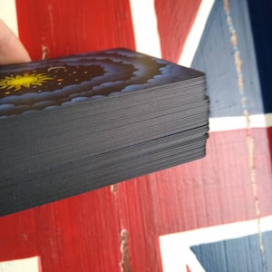 May include: A deck of cards with a black back and a colorful design on the front. The design features a yellow sun, blue clouds, and a starry night sky.