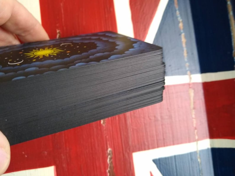 May include: A deck of cards with a black back and a colorful design on the front. The design features a yellow sun, blue clouds, and a starry night sky.