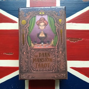 May include: A box for The Dark Mansion Tarot deck. The box is brown with gold accents and features an illustration of a woman in a black dress and a purple room with green curtains. The text "The Dark Mansion Tarot" is written in gold lettering.