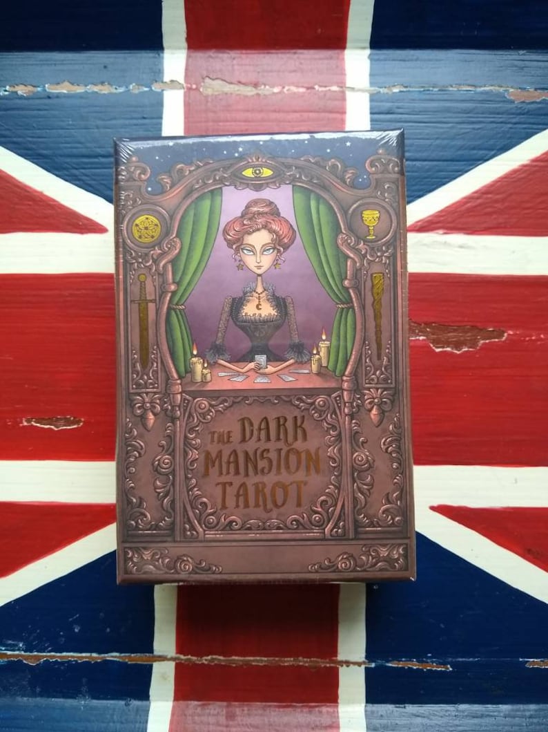 May include: A box for The Dark Mansion Tarot deck. The box is brown with gold accents and features an illustration of a woman in a black dress and a purple room with green curtains. The text "The Dark Mansion Tarot" is written in gold lettering.