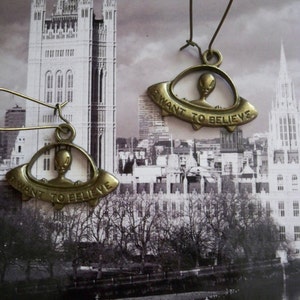 May include: A pair of bronze-colored earrings featuring a UFO design with an alien figure inside. The earrings have a hook closure and the UFOs have the text "I WANT TO BELIEVE" on them.