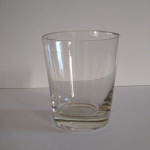 May include: A clear, cylindrical drinking glass with a thick, flat base. The glass has a slightly tapered shape, widening towards the top. The glass is empty and sits on a white surface.