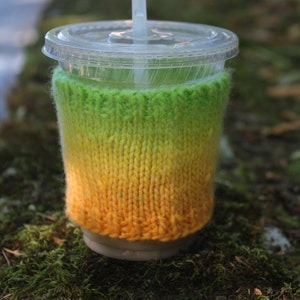 Può includere: Un bicchiere di plastica trasparente con un rivestimento in maglia verde, giallo e arancione. Il rivestimento ha un motivo sfumato. Una cannuccia di plastica trasparente sporge dalla parte superiore del bicchiere.