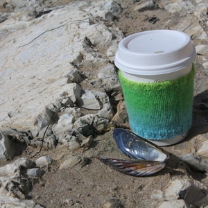 Pode incluir: Uma caneca de café branca com um aconchegante de malha verde e azul degradê em uma superfície rochosa. Há duas conchas marinhas azul-escuras ao lado da caneca.