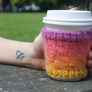 May include: A knitted coffee cup cozy with a gradient of purple, orange, and yellow. The cozy is on a white coffee cup.