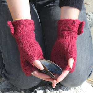 May include: A pair of red fingerless gloves with a fold-over cuff. The gloves are made of a soft, knitted fabric and have a cable knit pattern. The gloves are being held by a person, who is also holding a seashell.