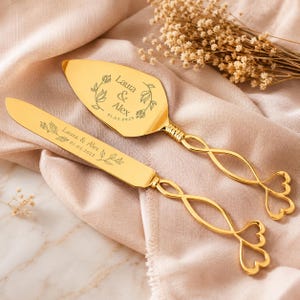 May include: A gold-toned cake knife and server set featuring twisted handles with heart-shaped ends. The blades are engraved with the names Laura & Alex, the date 01.05.2025, and floral accents. The metal surfaces have a polished, reflective finish.