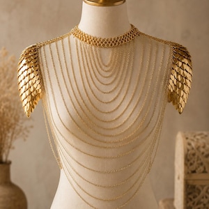 Puede incluir: Un collar de cuerpo en tono dorado con múltiples capas de cadenas finas drapeadas. La pieza incluye una gargantilla texturizada y dos hombreras construidas con placas metálicas doradas superpuestas en forma de escamas.
