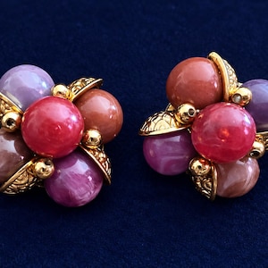 May include: A pair of clip-on earrings featuring a cluster of round, multi-coloured beads in shades of pink, purple, and coral. The beads are arranged around a central red sphere and accented with gold-toned metal components with textured details.