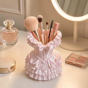 May include: A light pink makeup brush holder shaped like a corset with ruffled layers and a central bow. The holder contains several makeup brushes with various bristle types and a pink cosmetic tube, standing upright in the center of the structure.