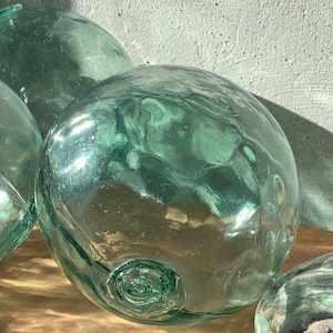 Può includere: Diversi grandi galleggianti rotondi in vetro trasparente con una leggera sfumatura verde acqua. I galleggianti hanno una superficie strutturata e un'indentazione circolare su un lato. L'immagine mette in risalto la trasparenza del vetro e le ombre.