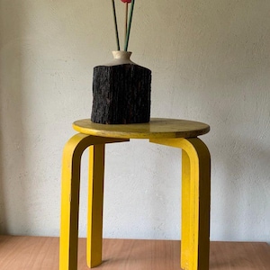 Könnte beinhalten: Ein gelber Beistelltisch aus Holz mit runder Platte und gebogenen Beinen. Eine dunkle, strukturierte Vase enthält drei Holz-Tulpen in Rosa- und Rottönen. Der Tisch steht auf einer Holzoberfläche.