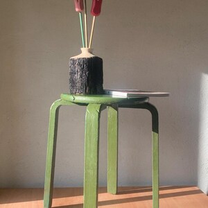 Könnte beinhalten: Ein grüner Holzstuhl trägt eine Vase mit drei Holz-Tulpen. Die Vase hat eine dunkle, strukturierte Außenseite und einen hellen Rand. Ein Buch liegt auf der Oberfläche des Hockers. Die Beine des Hockers sind gebogen.