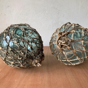 Puede incluir: Dos flotadores de pesca antiguos de vidrio con un tinte verde azulado, envueltos en una red protectora de cuerda natural. Los flotadores son esféricos y presentan un aspecto envejecido, indicando su antigüedad. Se encuentran sobre una superficie de madera.