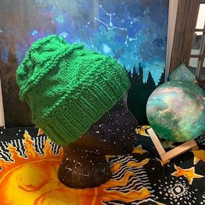 May include: A green knitted beanie hat with a cable knit pattern. The hat is displayed on a mannequin head against a colorful background.