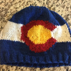 May include: Knitted hat featuring the Colorado state flag. The hat is blue, white, red, and yellow.