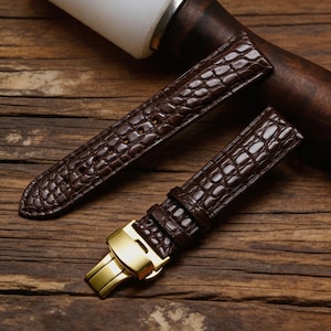 Pode incluir: Duas pulseiras de relógio castanhas em couro com padrão de pele de crocodilo. As pulseiras têm um fecho dourado. As pulseiras de relógio são apresentadas numa superfície de madeira.