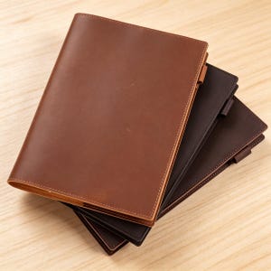 May include: Three leather journals in shades of brown, stacked on a light wood surface. The top journal is a light brown, the middle is a dark brown, and the bottom is a deep brown. Each journal has a tab.