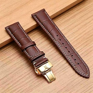 May include: A two-piece dark brown watch strap featuring a textured alligator-style embossed pattern, white contrast stitching along the edges, and a polished gold-tone metal butterfly deployment clasp with a secure locking mechanism.