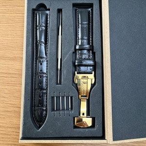 Pode incluir: Uma pulseira de relógio em couro preto com textura de crocodilo, apresentando um fecho de báscula em metal dourado. O conjunto inclui uma ferramenta para pinos de mola e quatro pequenos pinos metálicos, organizados numa caixa retangular.