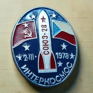 May include: An oval metal pin featuring a central white rocket with red accents and the text SOYUZ-28. The blue background displays the Soviet flag, the Czechoslovakian flag, white stars, the date 2.III.1978, and the Cyrillic text INTERKOSMOS.