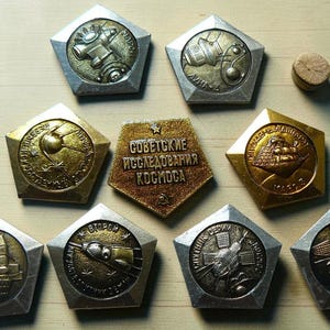 May include: A collection of nine pentagonal metal pins featuring space exploration themes. Eight pins display relief images of spacecraft and satellites, while the central pin is gold-toned with the Cyrillic text "СОВЕТСКИЕ ИССЛЕДОВАНИЯ КОСМОСА".