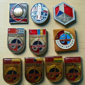 May include: A collection of eleven metal enamel pins featuring Interkosmos space program themes. The pins display various geometric shapes, national flags, and Cyrillic text including CCCP, Interkosmos, and dates like 1978 and 1984, with red, blue, gold, and white colors.