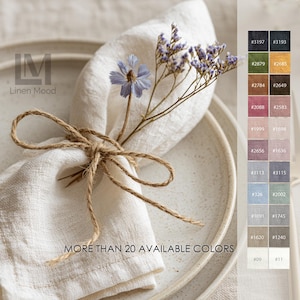 Peut inclure: Une serviette en lin blanc pliée sur une assiette en céramique mouchetée, nouée avec un ruban en jute naturel et décorée d'un petit brin de fleurs violettes séchées. L'image inclut une palette de couleurs avec le texte MORE THAN 20 AVAILABLE COLORS.