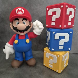 Könnte beinhalten: Eine Mario-Figur mit roter Kappe mit weißem "M", rotem Hemd, blauer Latzhose und weißen Handschuhen steht neben drei gestapelten Fragezeichen-Blöcken in Rot, Blau und Gelb.