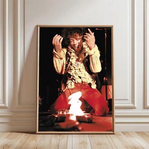 May include: Framed photograph of a live performance. The subject is kneeling, wearing a yellow shirt with a ruffled collar and red trousers. A small fire is in front of the subject. The image is in a gold frame.