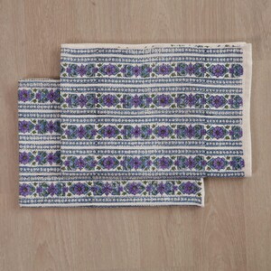 May include: A rectangular textile featuring a repeating pattern of purple floral motifs and blue geometric shapes arranged in horizontal bands. The design includes thin white and blue striped borders separating the floral rows on a light background.