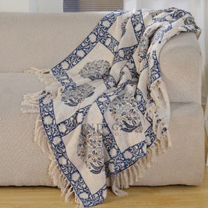 May include: A cream-coloured throw blanket with a blue floral pattern and fringe detail draped over a beige sofa. The blanket features a repeating square design with floral motifs and decorative borders. The fringe is a light beige colour.