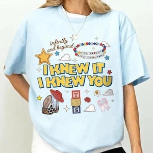 May include: A light blue t-shirt featuring a graphic print with the text "infinity and beyond", "I KNEW IT I KNEW YOU", and "YOU'VE GOT A FRIEND IN ME". The design includes a yellow star, a cowboy boot, alphabet blocks, and various small icons.
