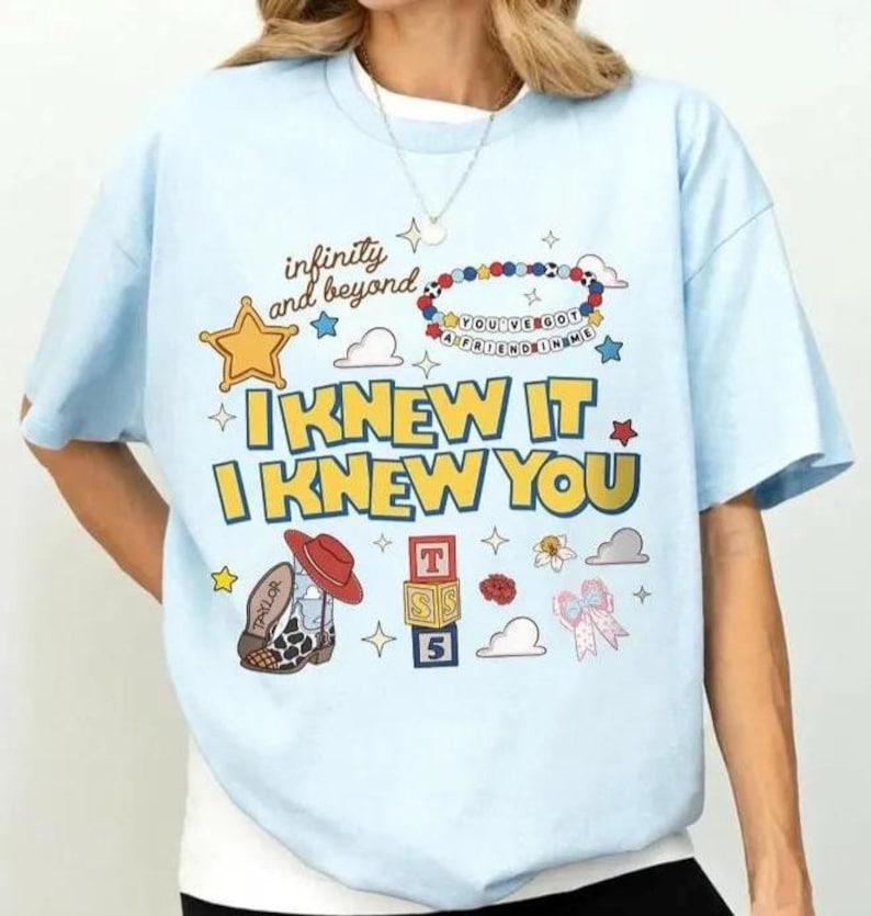 May include: A light blue t-shirt featuring a graphic print with the text "infinity and beyond", "I KNEW IT I KNEW YOU", and "YOU'VE GOT A FRIEND IN ME". The design includes a yellow star, a cowboy boot, alphabet blocks, and various small icons.