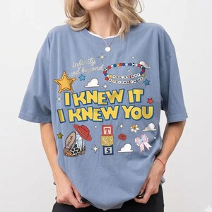 May include: A dusty blue t-shirt featuring a graphic print with the text "infinity and beyond," "YOU'VE GOT A FRIEND IN ME," "I KNEW IT," and "I KNEW YOU." The design includes a yellow star, a beaded bracelet, a cowboy boot, alphabet blocks, and a bow.