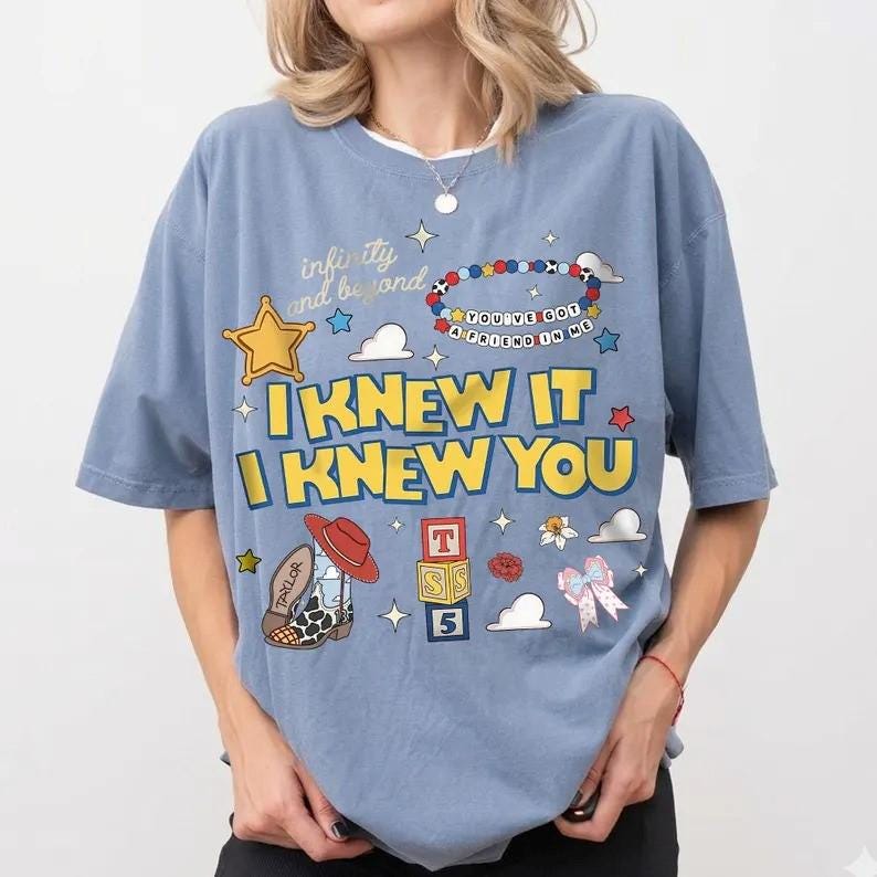 May include: A dusty blue t-shirt featuring a graphic print with the text "infinity and beyond," "YOU'VE GOT A FRIEND IN ME," "I KNEW IT," and "I KNEW YOU." The design includes a yellow star, a beaded bracelet, a cowboy boot, alphabet blocks, and a bow.