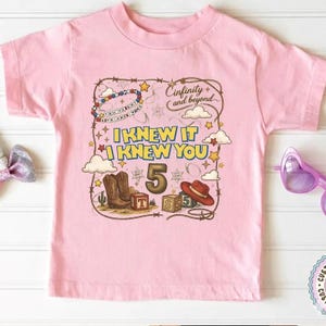 May include: A light pink short-sleeve t-shirt featuring a graphic print with the text "Cinfinity and beyond", "I KNEW IT I KNEW YOU", and the number "5". The design includes illustrations of cowboy boots, a hat, blocks, and stars.