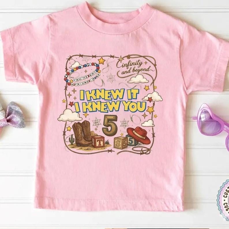 May include: A light pink short-sleeve t-shirt featuring a graphic print with the text "Cinfinity and beyond", "I KNEW IT I KNEW YOU", and the number "5". The design includes illustrations of cowboy boots, a hat, blocks, and stars.