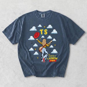 May include: A dark blue short-sleeve t-shirt featuring a graphic of a person with long hair wearing a yellow plaid shirt and cow-print pants, holding a guitar. The design includes white clouds, yellow stars, a red hat, and the text TS I KNEW IT I KNEW YOU.
