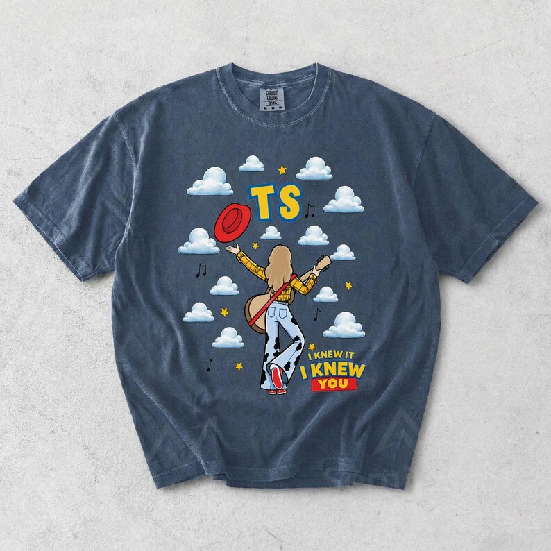 May include: A dark blue short-sleeve t-shirt featuring a graphic of a person with long hair wearing a yellow plaid shirt and cow-print pants, holding a guitar. The design includes white clouds, yellow stars, a red hat, and the text TS I KNEW IT I KNEW YOU.