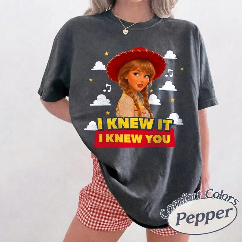 May include: A charcoal gray short-sleeve t-shirt featuring a graphic of a cartoon character with braided hair wearing a red cowboy hat. The design includes white clouds, yellow stars, musical notes, and the text I KNEW IT I KNEW YOU in yellow and red.
