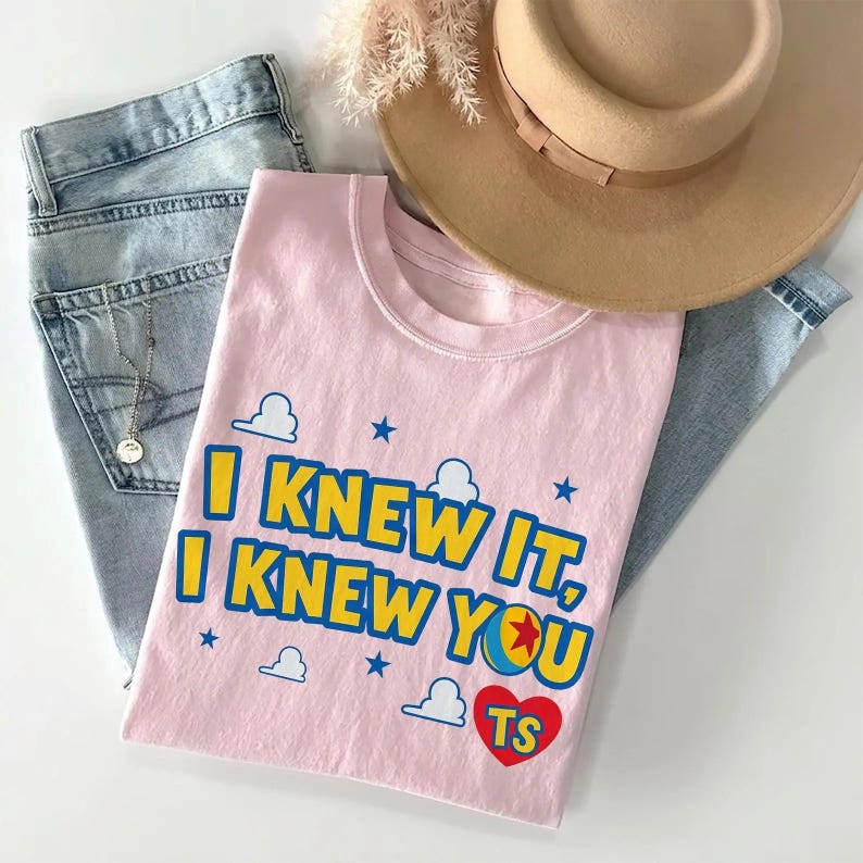 May include: A light pink t-shirt featuring the blue and yellow text "I KNEW IT, I KNEW YOU" with a red heart containing the letters "TS" and a small ball graphic. The shirt is displayed alongside a pair of light blue denim jeans and a tan hat.