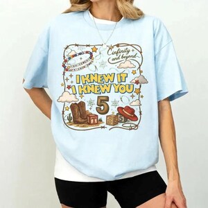 May include: A light blue t-shirt featuring a graphic print with a lasso border, cowboy boots, a red hat, and wooden blocks. The text reads "I KNEW IT I KNEW YOU", "5", and "Cinfinity and beyond". The design includes stars, clouds, and a beaded string.