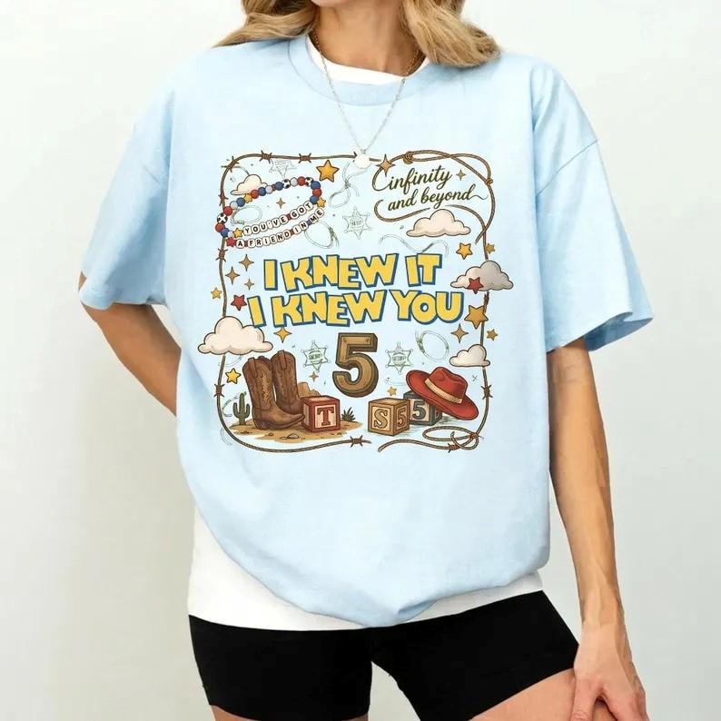 May include: A light blue t-shirt featuring a graphic print with a lasso border, cowboy boots, a red hat, and wooden blocks. The text reads "I KNEW IT I KNEW YOU", "5", and "Cinfinity and beyond". The design includes stars, clouds, and a beaded string.