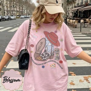 May include: A light pink t-shirt featuring a graphic print of a cowboy boot with a cow print pattern, a red hat, and blocks spelling TS. The boot has the name Taylor written on it. A black quilted shoulder bag with a chain strap is worn over the shoulder.